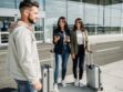 Dve žene sa koferima stoje ispred aerodroma i razgovaraju sa muškarcem