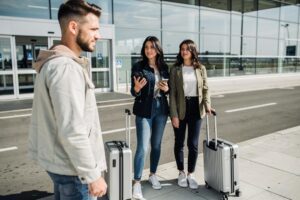 Dve žene sa koferima stoje ispred aerodroma i razgovaraju sa muškarcem