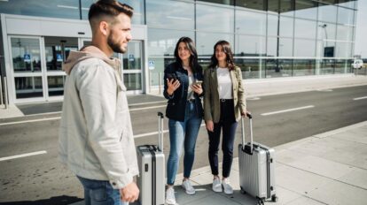 Dve žene sa koferima stoje ispred aerodroma i razgovaraju sa muškarcem