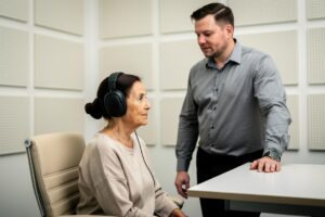 Starija žena sa slušalicama sedi u sobi sa zvučnim panelima dok muškarac stoji pored stola